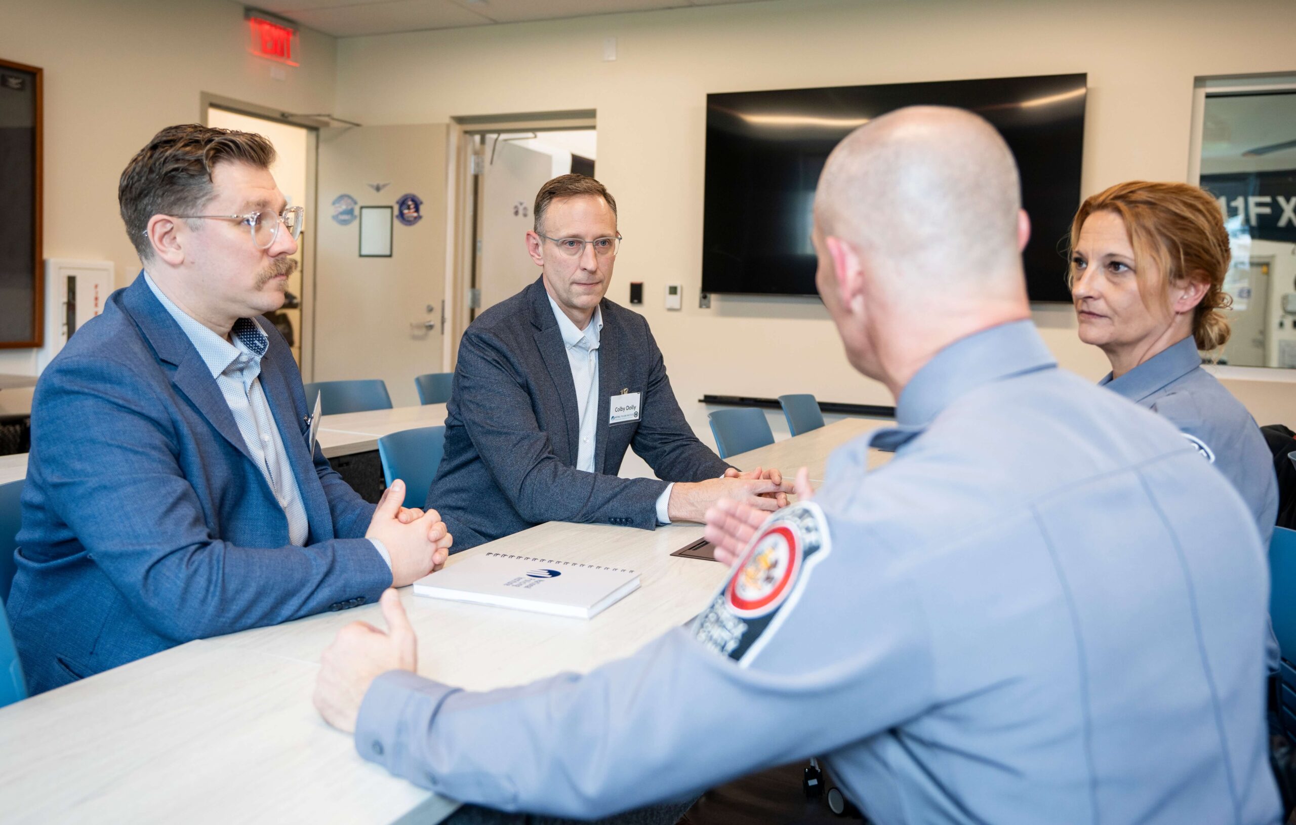 Two consultants meet with two uniformed personnel in a classroom-style setting to discuss public safety initiatives.