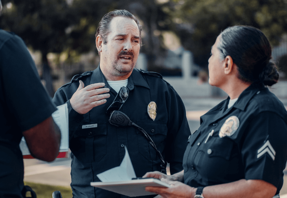 Two uniformed police officers discuss information in an outdoor setting while reviewing a notepad.