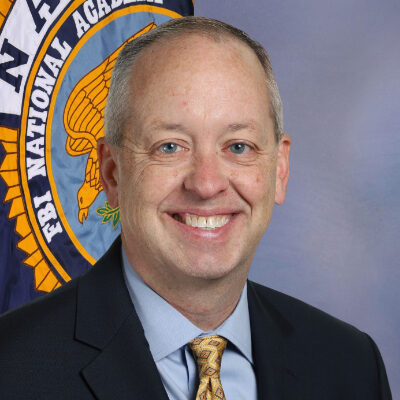 A smiling man in a suit stands in front of an FBI National Academy flag.