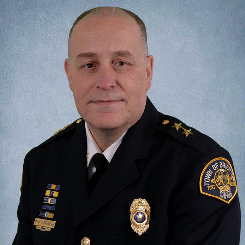 Portrait of Chief David Catholdi A man in a police dress uniform with badges and insignia sits in front of a blue textured background. His uniform patch reads “Town of Brighton Police.