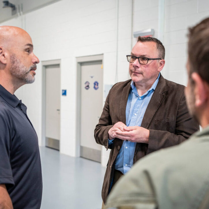 Three men stand in conversation inside a facility with white walls and closed doors in the background.