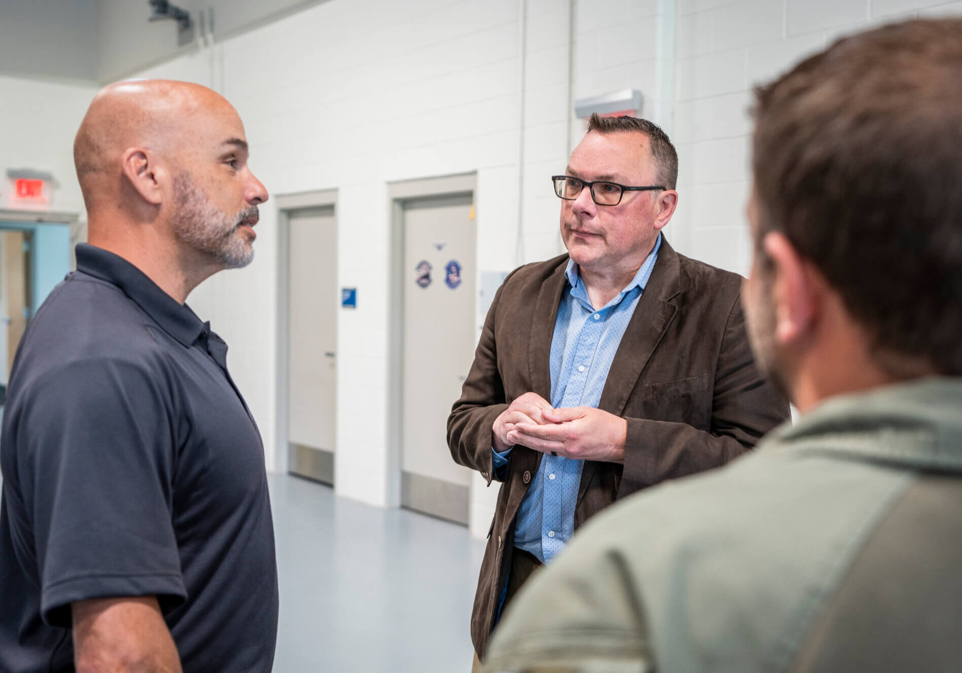 Professionals Discuss in Hallway Three men stand in conversation inside a facility with white walls and closed doors in the background.